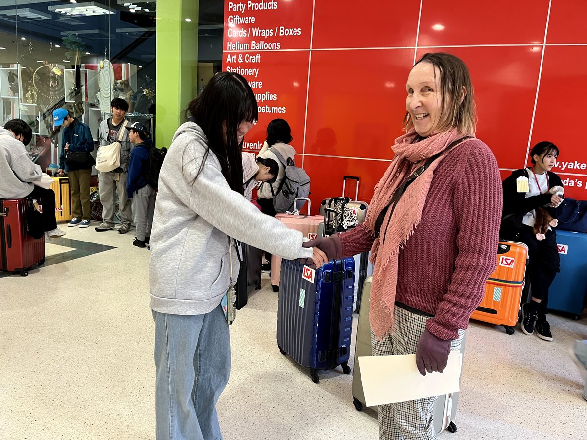 A warm, candid arrival scene showing a New Zealand host or support person shaking hands with an arriving...