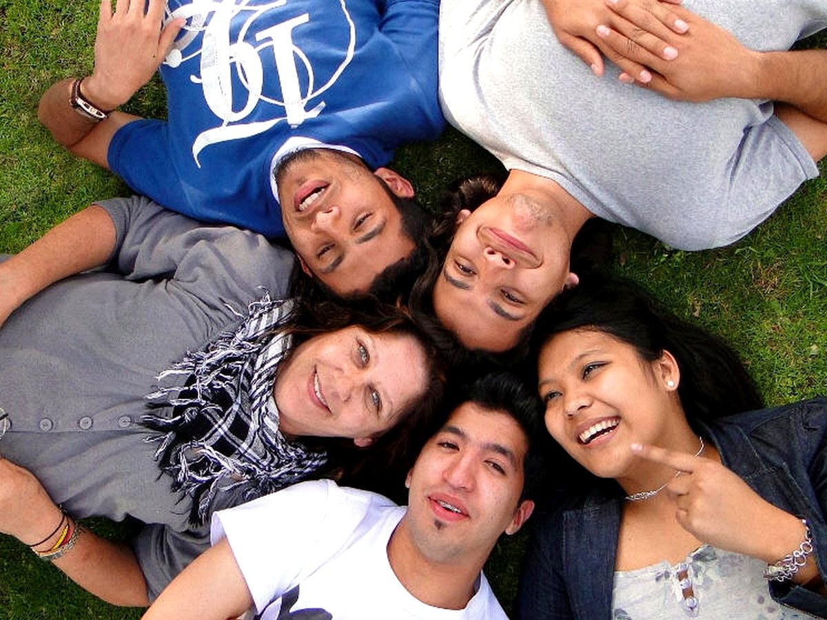 An overhead bird's-eye view of five ethnically diverse students lying in a circle on green grass with...