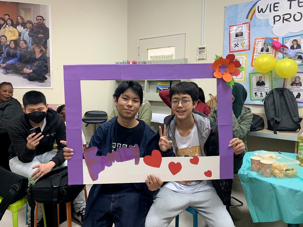 Two young male students pose happily inside a handcrafted purple 'FAMILY' photo frame prop during a school...