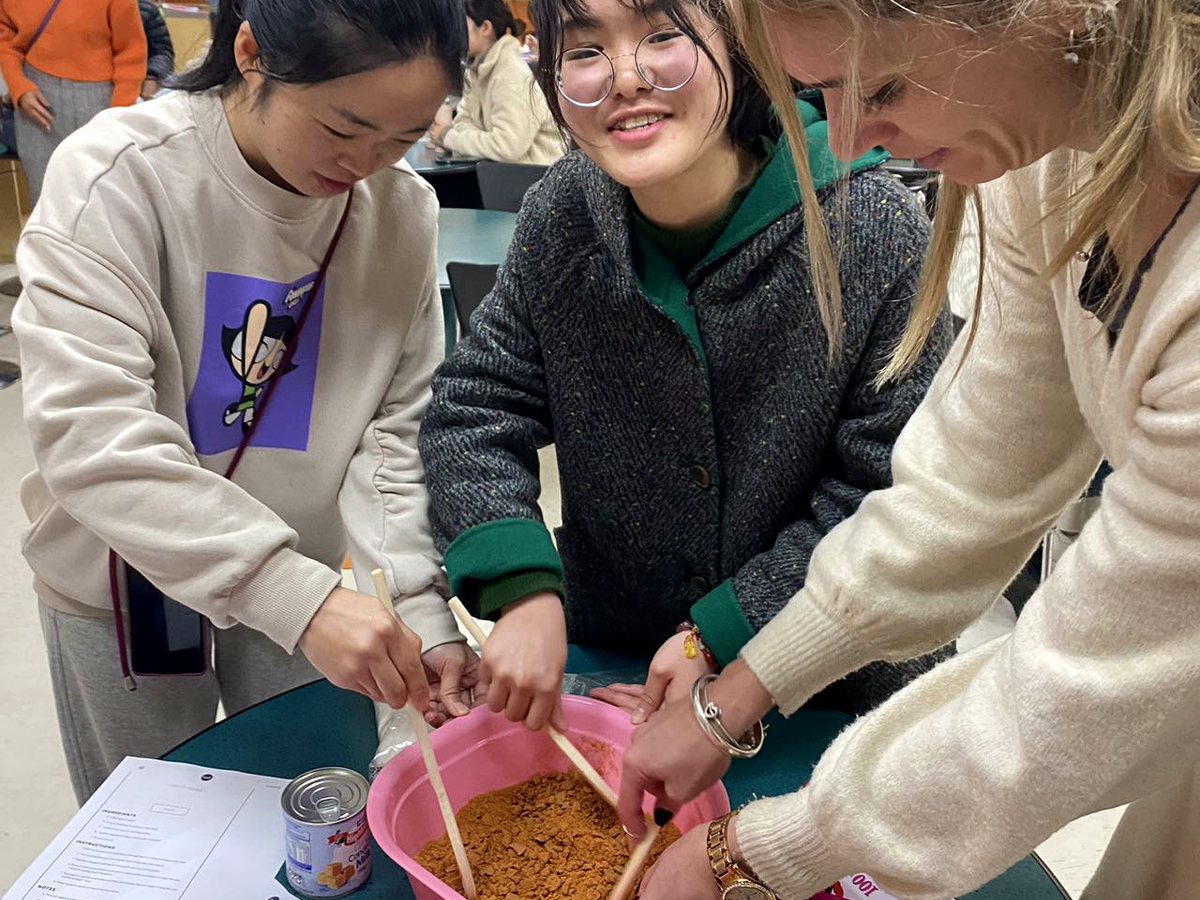 Three female international students actively engaged in a hands-on New Zealand cultural cooking activity,...