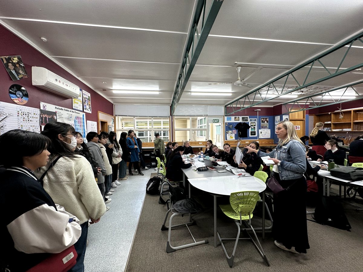 An authentic, active classroom scene showing a teacher in a denim jacket addressing a table of seated...